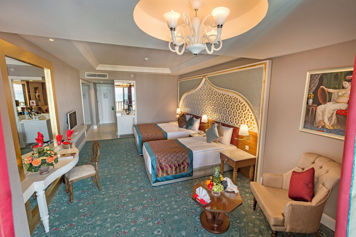 A hotel room with two beds, a sitting area with a chair and a table, a mirror, and a chandelier.
