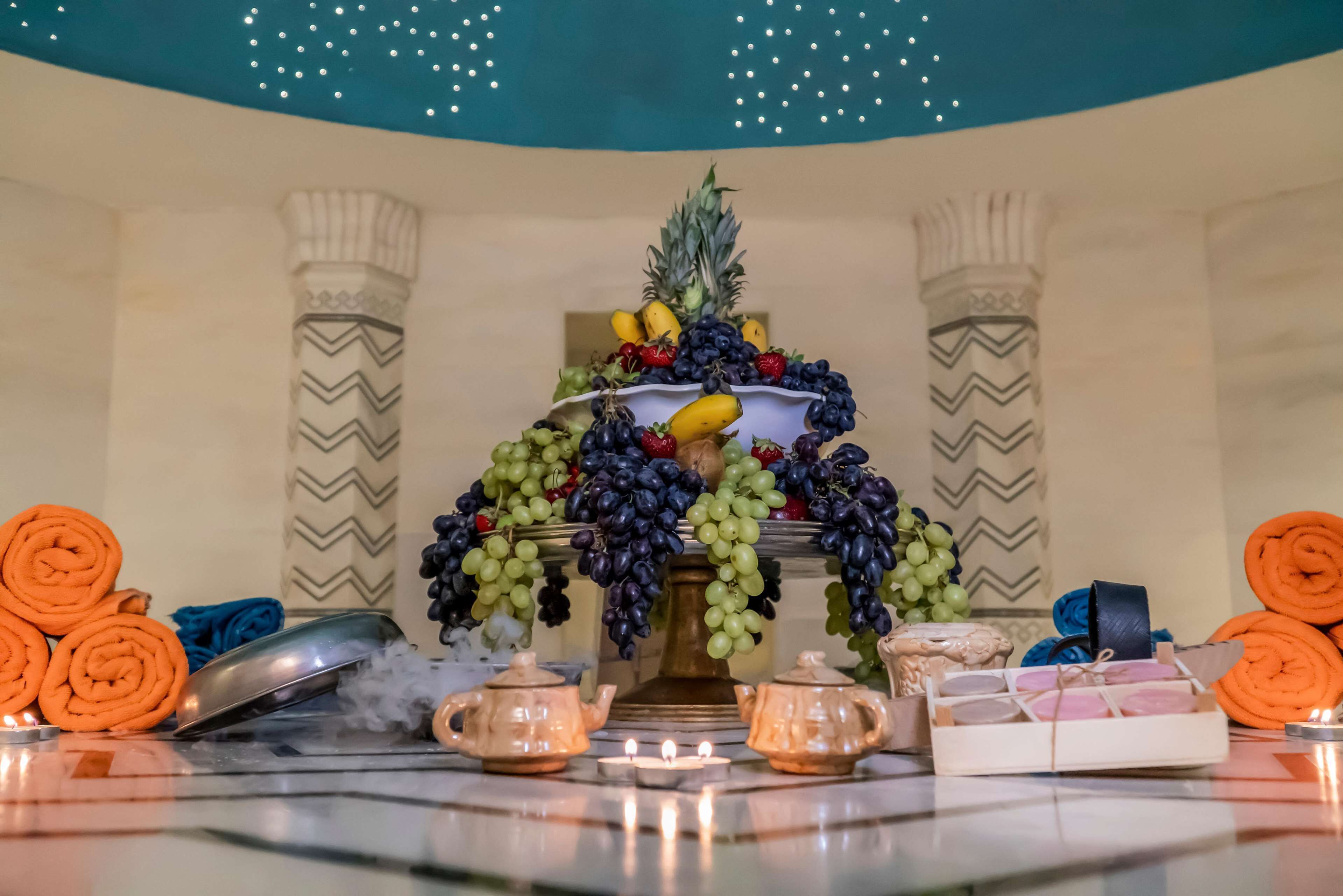 A decorative fruit arrangement on a table with candles, rolled towels, and small containers.
