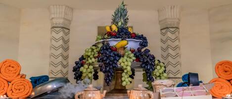 A decorative fruit arrangement on a table with candles, rolled towels, and small containers.