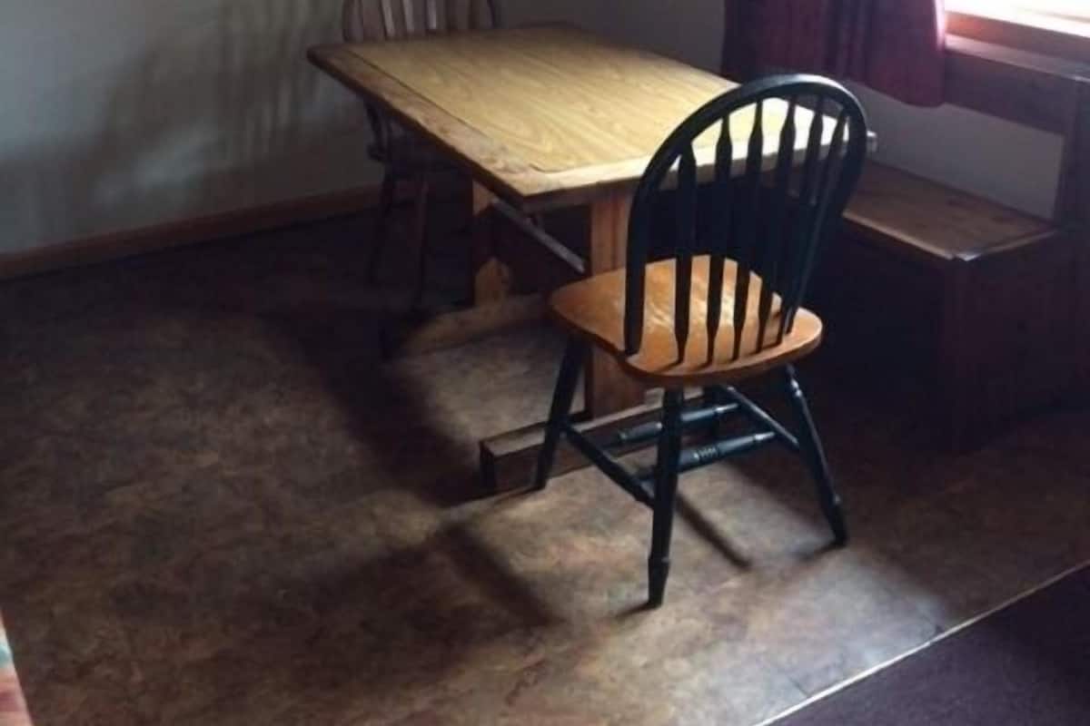 A dining area with a wooden table and four chairs, a hanging light fixture, a framed picture on the wall, and a window with curtains.