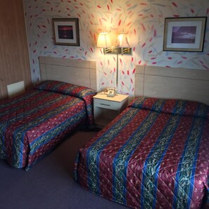 A hotel room with two beds, each with a patterned bedspread, a bedside table with a lamp, and a wall with a floral wallpaper design.