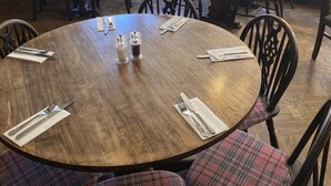 A dining area with round tables and chairs, large windows with curtains, and a radiator. - The New Inn Hotel (Lechlade)