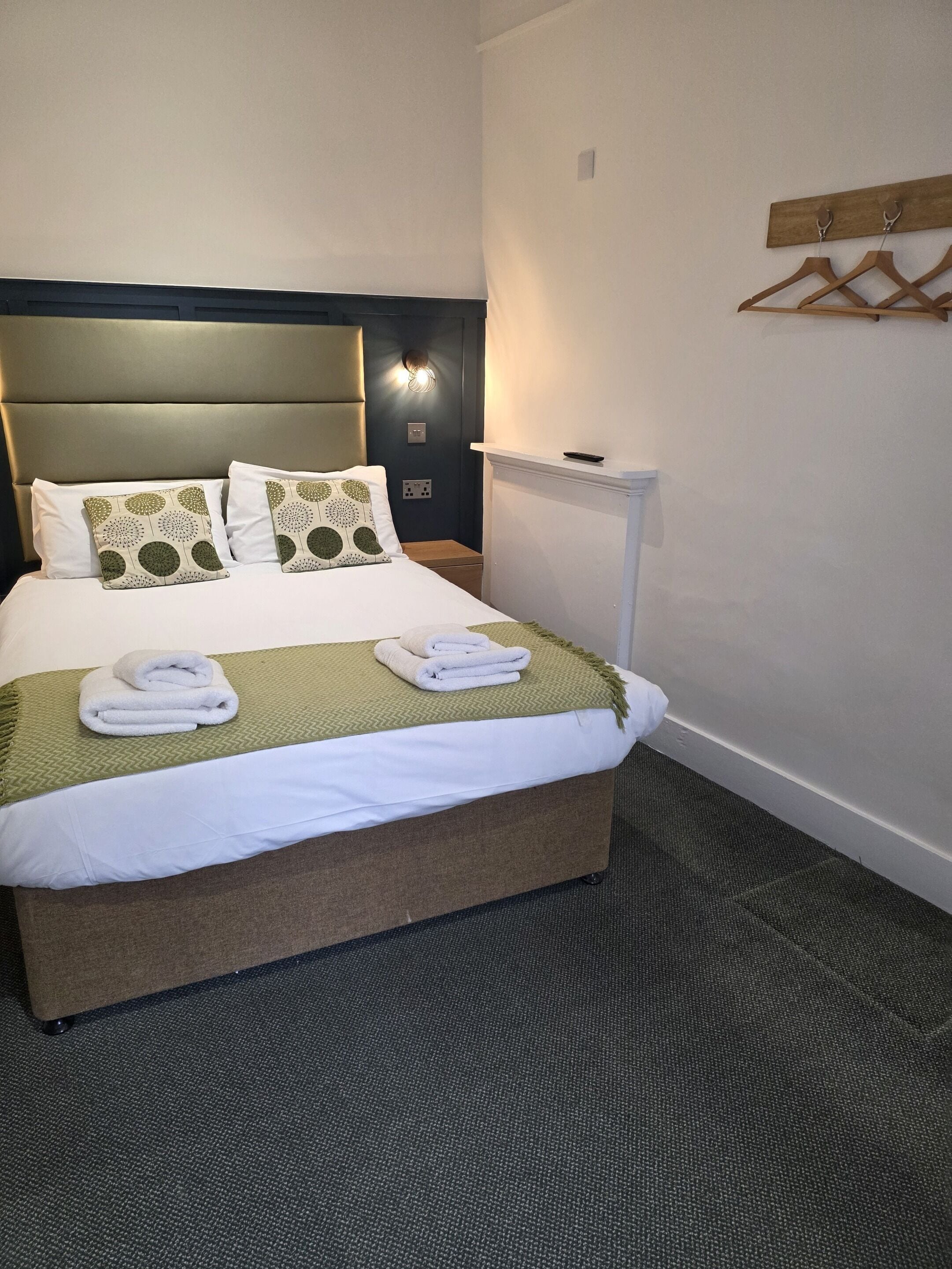A neatly made bed with two pillows, a bedside table with a remote control, and a wall-mounted coat rack.