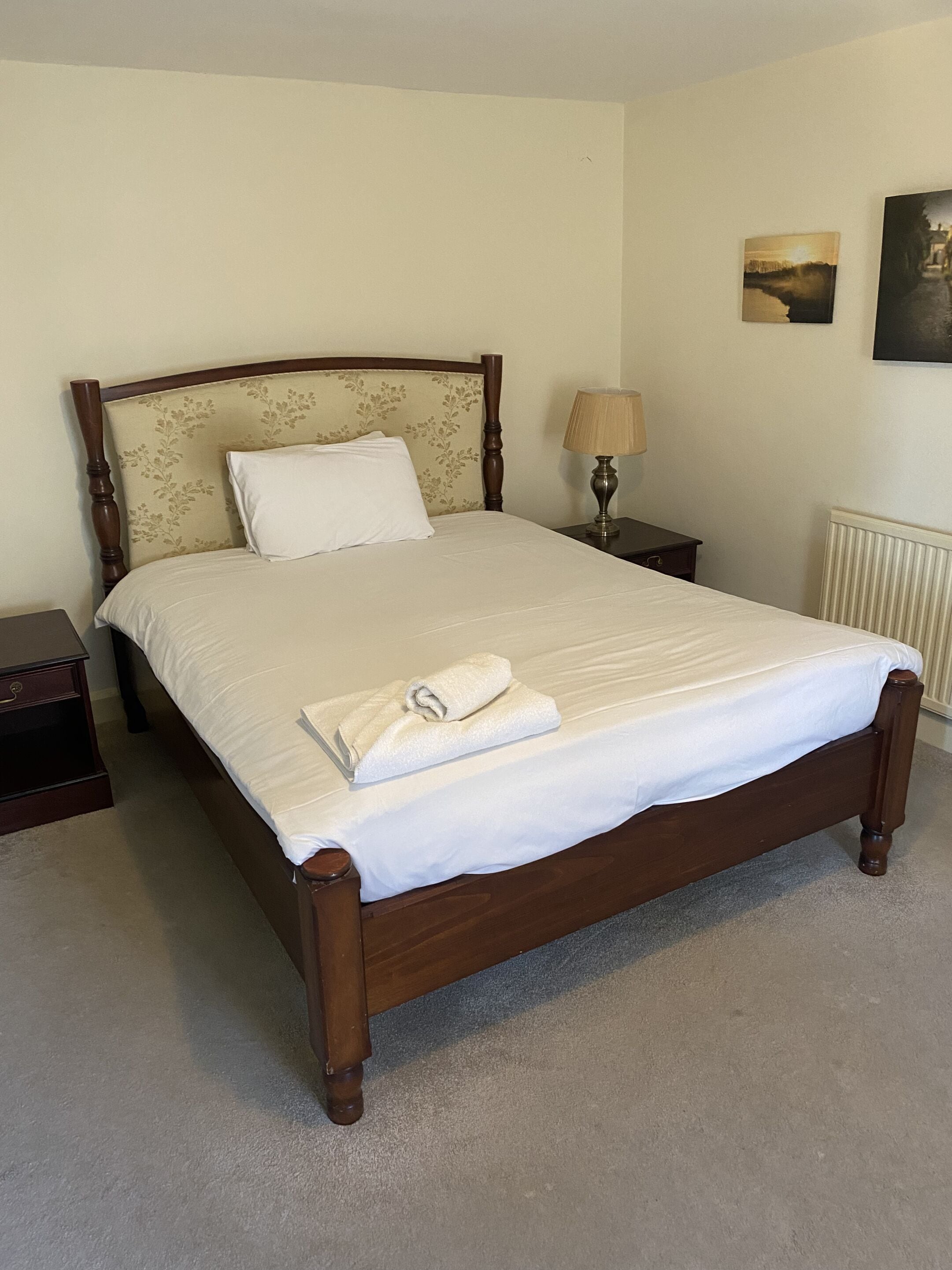 Un lit bien fait, avec une couette blanche et une serviette blanche enroulée, une tête de lit en bois ornée de motifs floraux, une lampe de chevet et un tableau encadré accroché au mur.