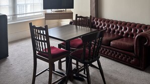 A room with a dining table, chairs, a sofa, a television, and a window with blinds. - The New Inn Hotel (Lechlade)