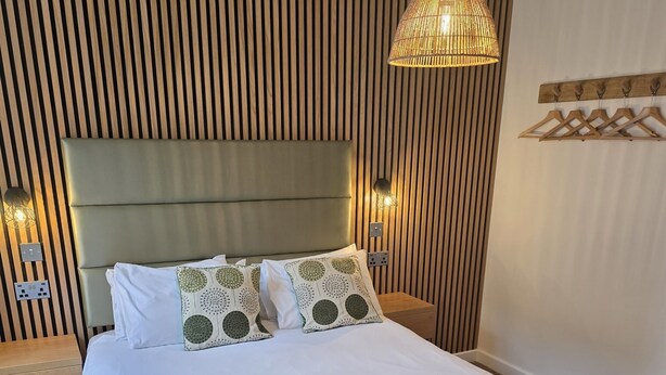 A neatly made bed with a green throw blanket and folded towels, a wooden slatted headboard, and a hanging light fixture. - The New Inn Hotel (Lechlade)