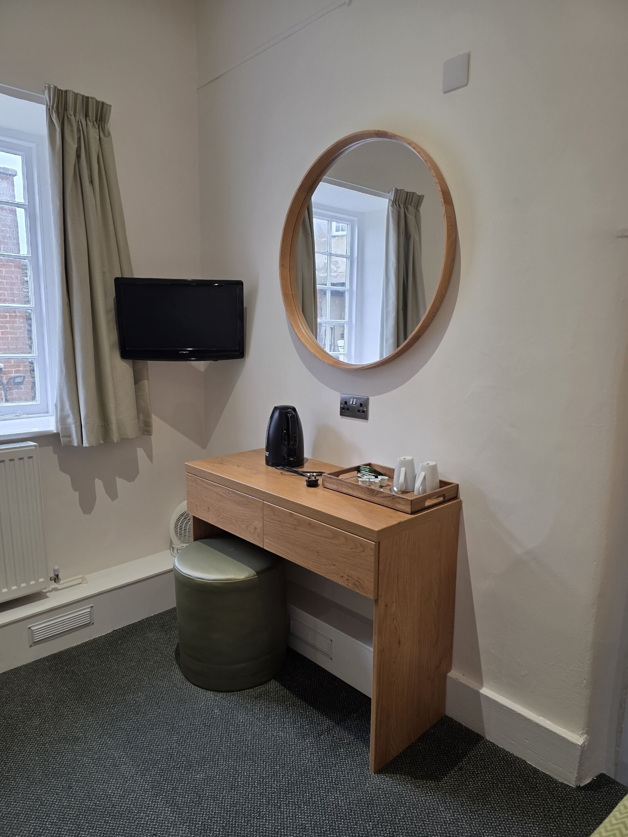 A hotel room with a wooden desk, a round mirror, a flat-screen TV, a radiator, a waste bin, and a window with curtains.