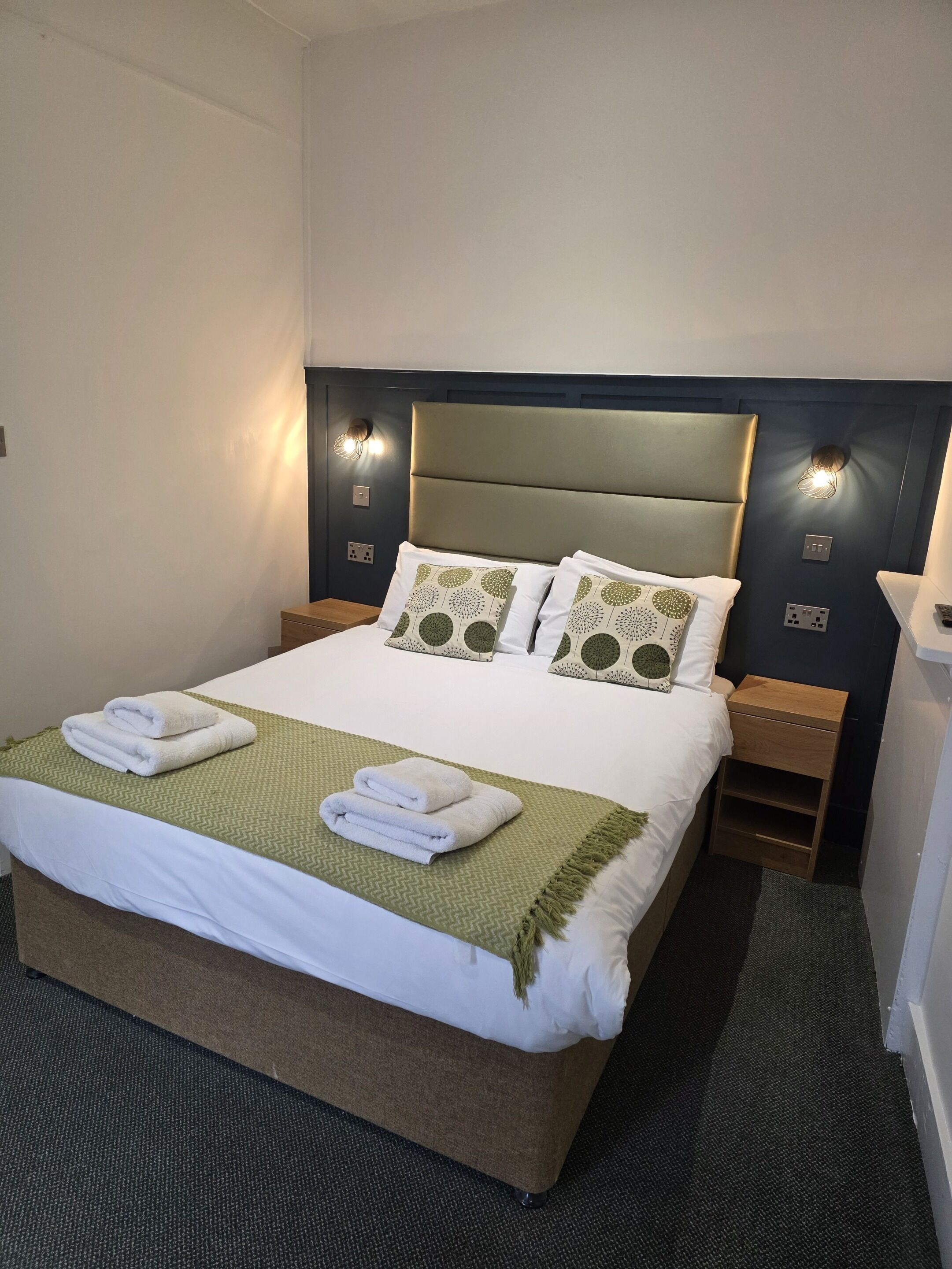 A neatly made bed with a green throw blanket and two folded towels, a headboard with built-in lighting, and bedside tables with lamps.