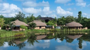 A series of thatched-roof cottages with a central archway, surrounded by lush greenery and a calm reflective water body. - Montis Resort (Pai)