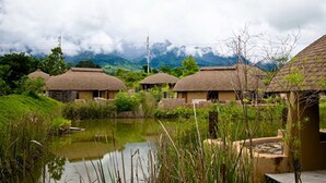 A row of thatched-roof cottages with a pond in the foreground, surrounded by lush greenery and mountains in the background. - Montis Resort (Pai)