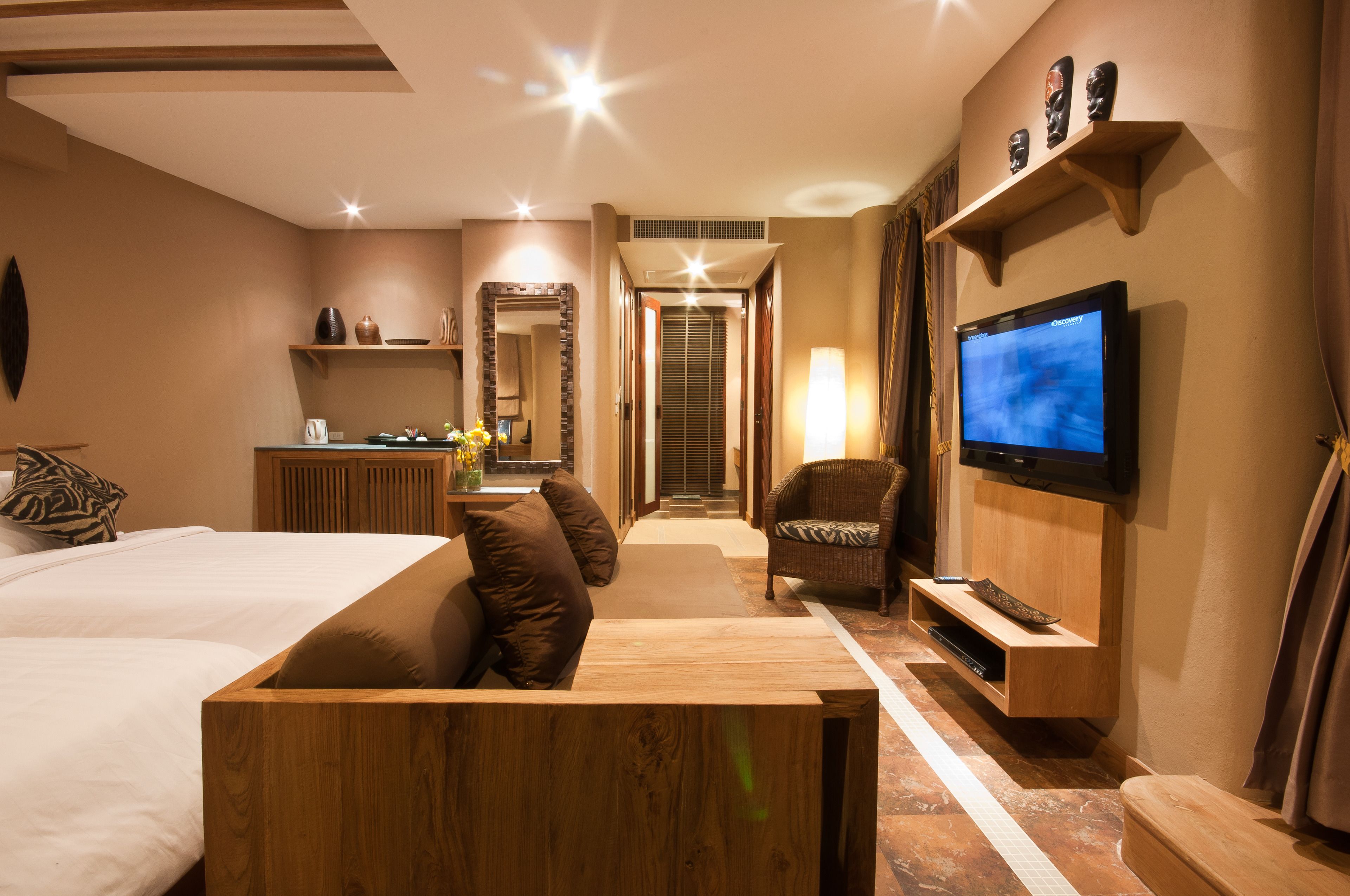 Habitación de hotel con cama, un banco de madera, televisión, espejo y estante con adornos.