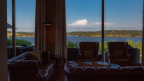 A room with a large window overlooking a body of water and greenery. There are leather sofas and armchairs arranged for seating. - Montargil Monte Novo (Ponte de Sor)