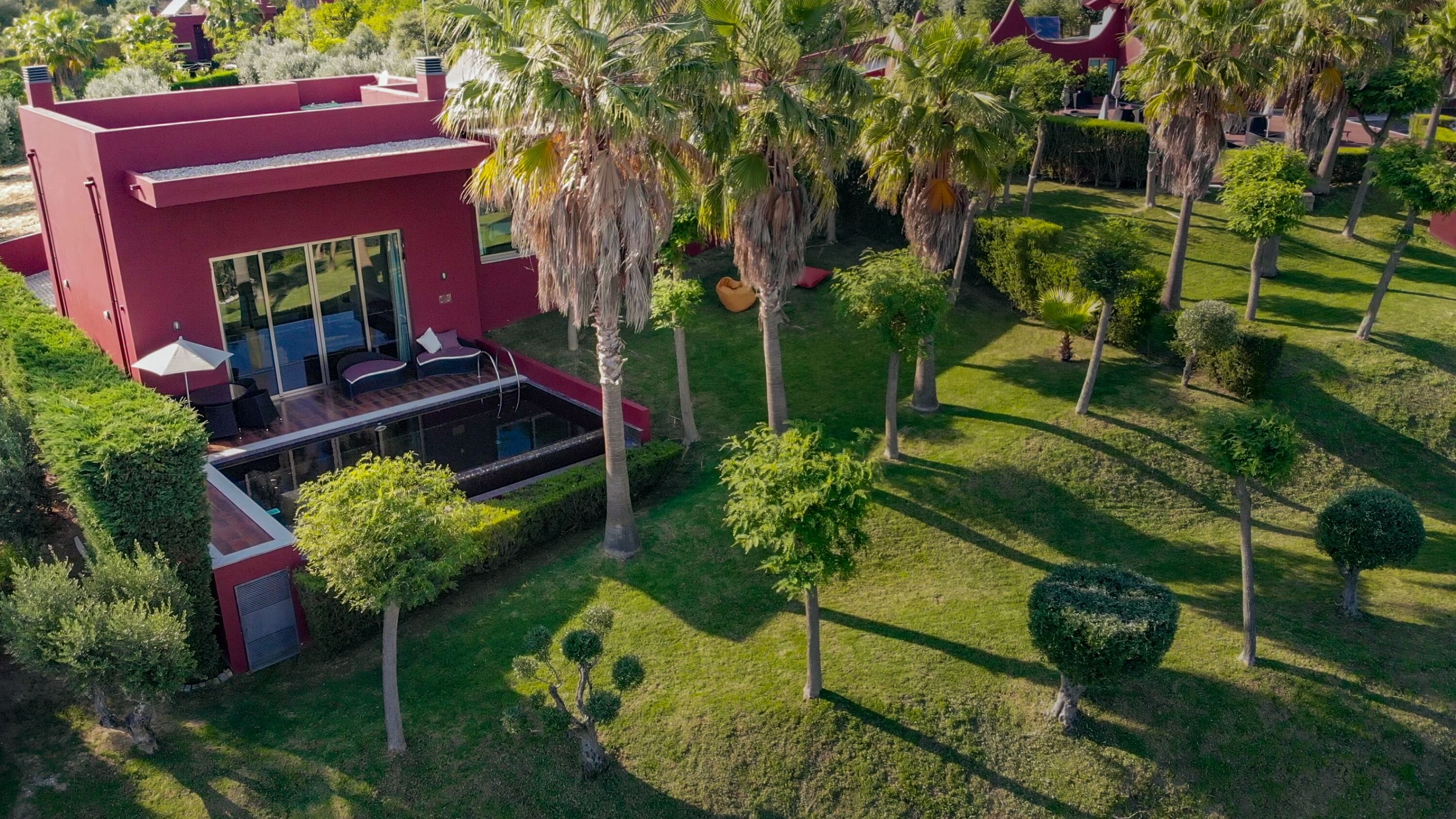 A modern building with a red facade, a pool area with lounge chairs, and a well-maintained garden with various trees and shrubs.
