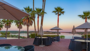 An outdoor patio with round tables and chairs, surrounded by palm trees and overlooking a body of water. - Montargil Monte Novo (Ponte de Sor)