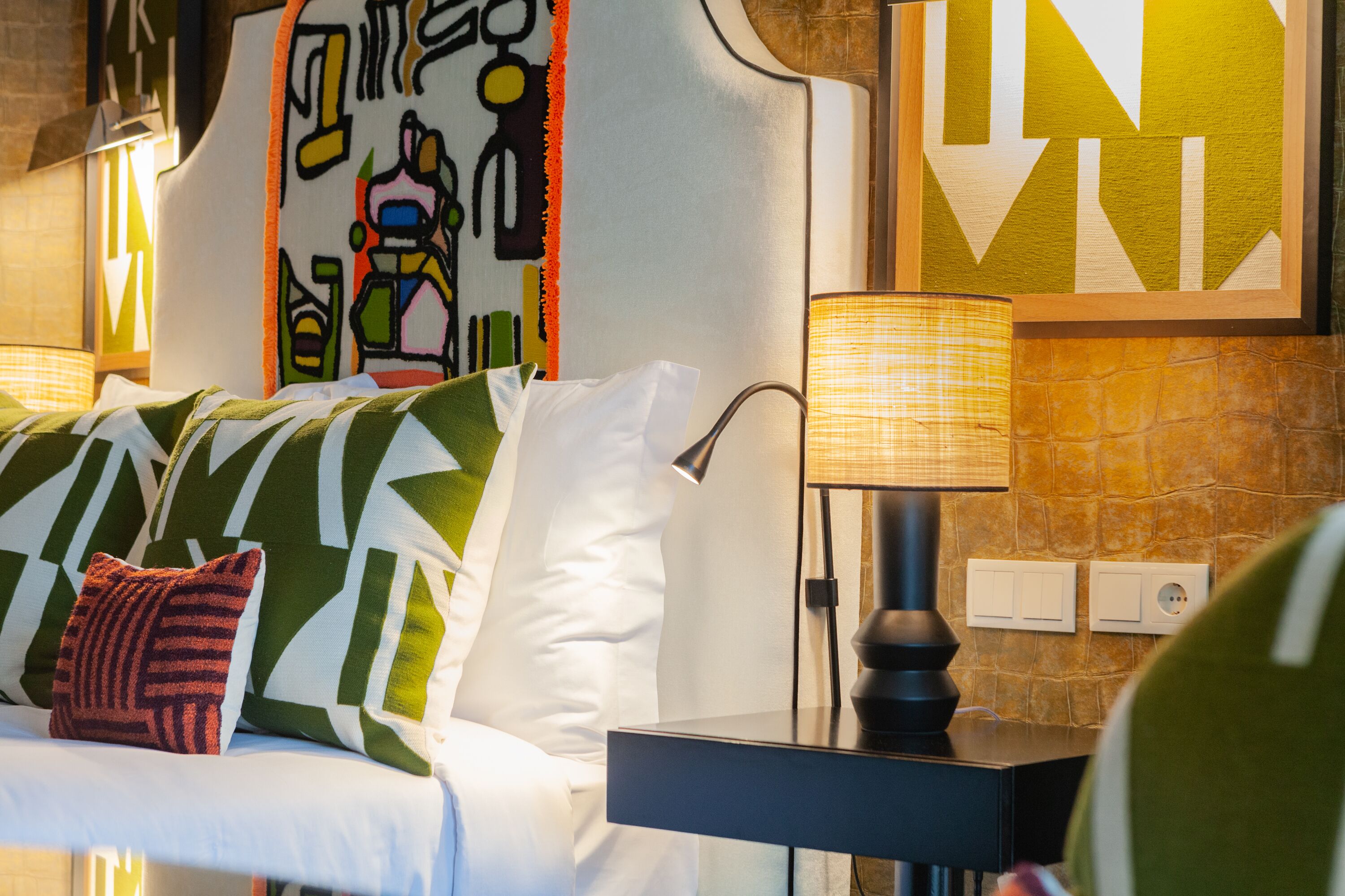 Una habitación de hotel moderna con un sofá de patrón verde y blanco, una mesita auxiliar negra con una lámpara y una obra de arte mural geométrica.