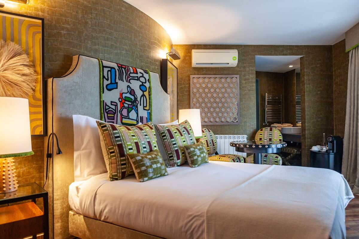 A hotel room with a large bed, a headboard with artwork, a round table with a lamp, and a built-in heater.
