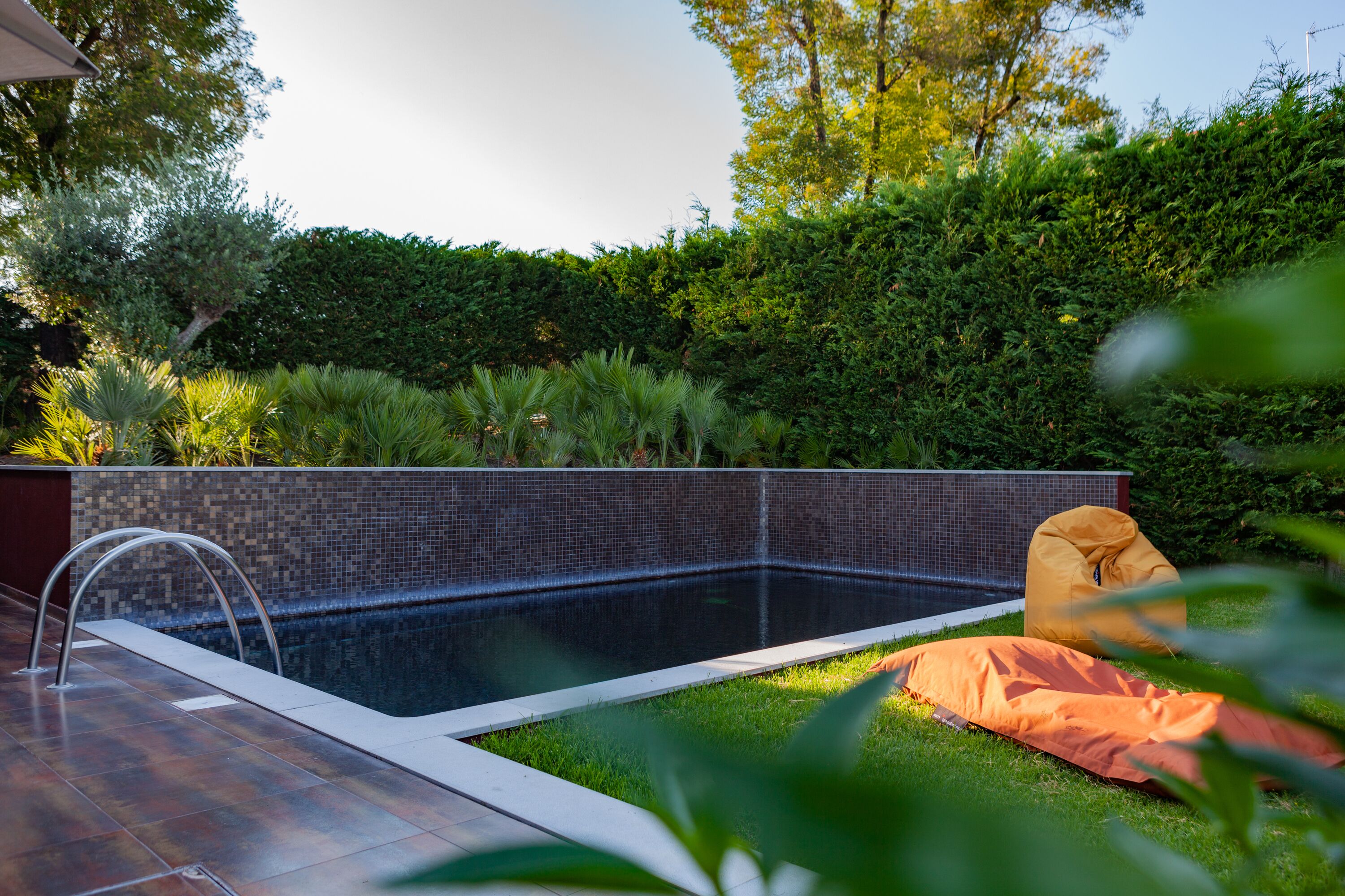 Piscina com borda azulejada, cercada por um conjunto de sebe bem cuidado e um gramado com uma poltrona de encosto.