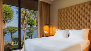 A large bed with a tufted headboard, a nightstand with a lamp, and a view of palm trees and water through floor-to-ceiling windows. - Montargil Monte Novo (Ponte de Sor)