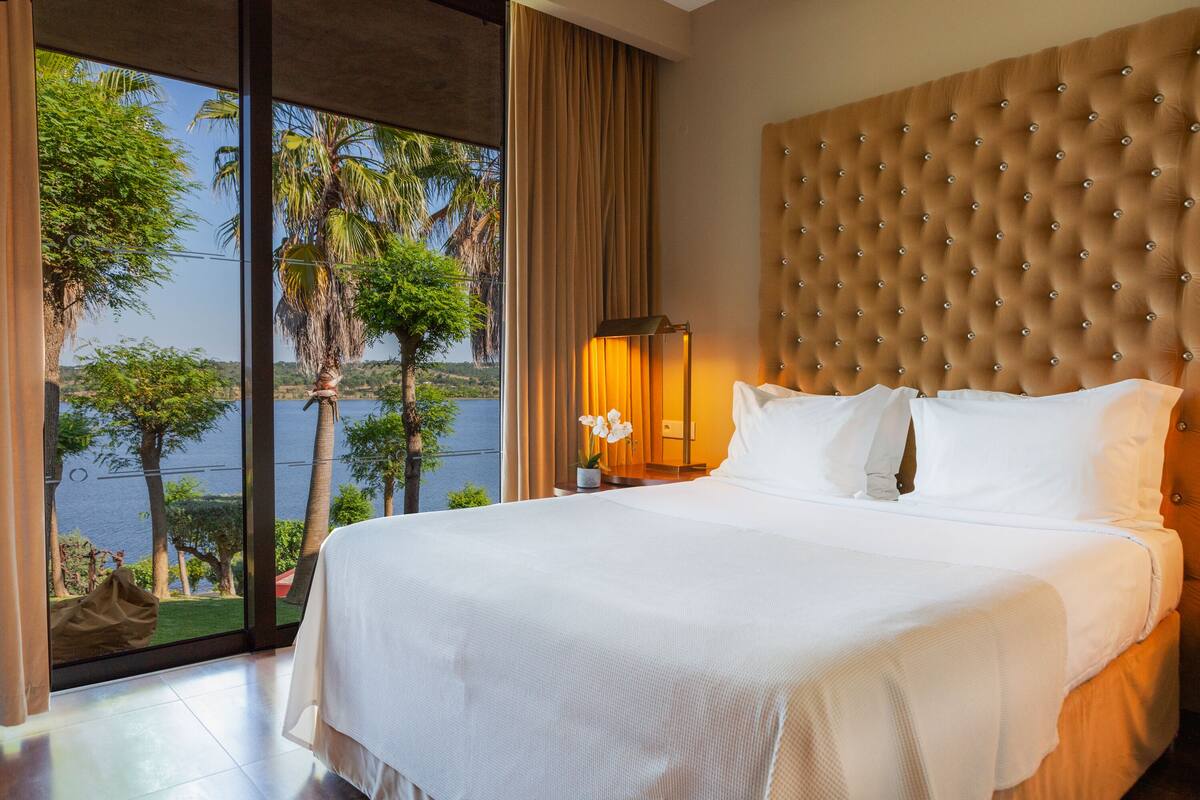 A large bed with a tufted headboard, a nightstand with a lamp, and a view of palm trees and water through floor-to-ceiling windows.