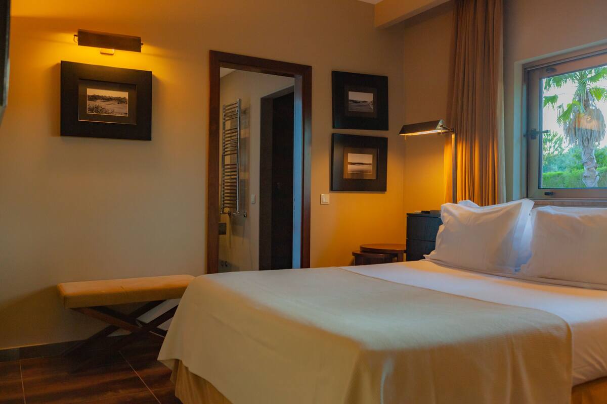 A hotel room with a bed, a bench, a small table, and framed pictures on the wall.