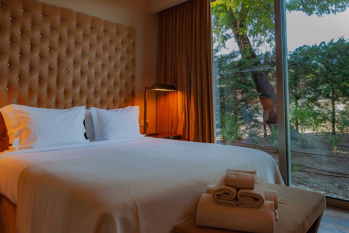 A hotel room with a tufted headboard, a large bed with white linens, a nightstand with a lamp, and a view of trees through the window.