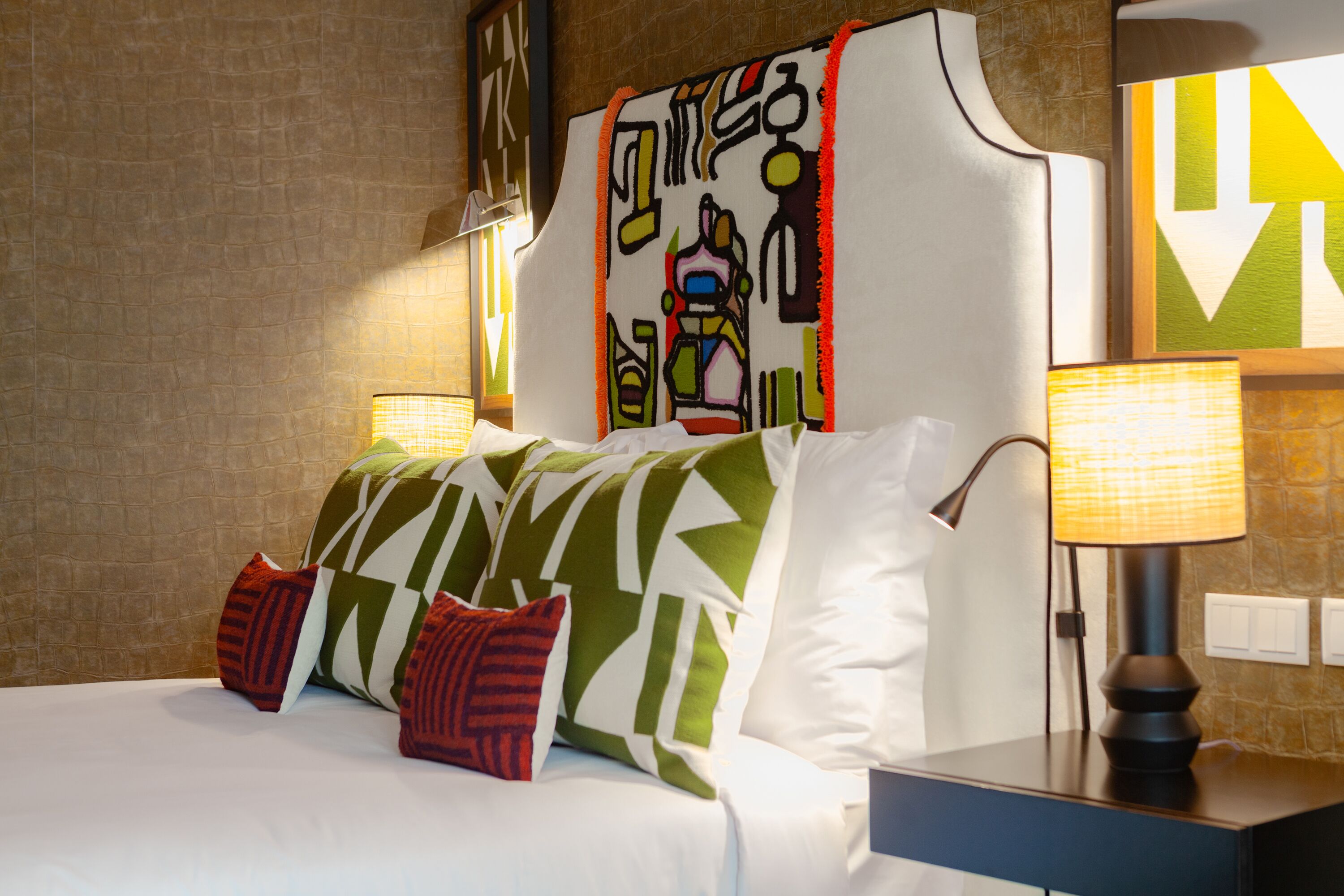 A hotel room with a bed, pillows, a nightstand, a lamp, and a wall with a geometric painting.