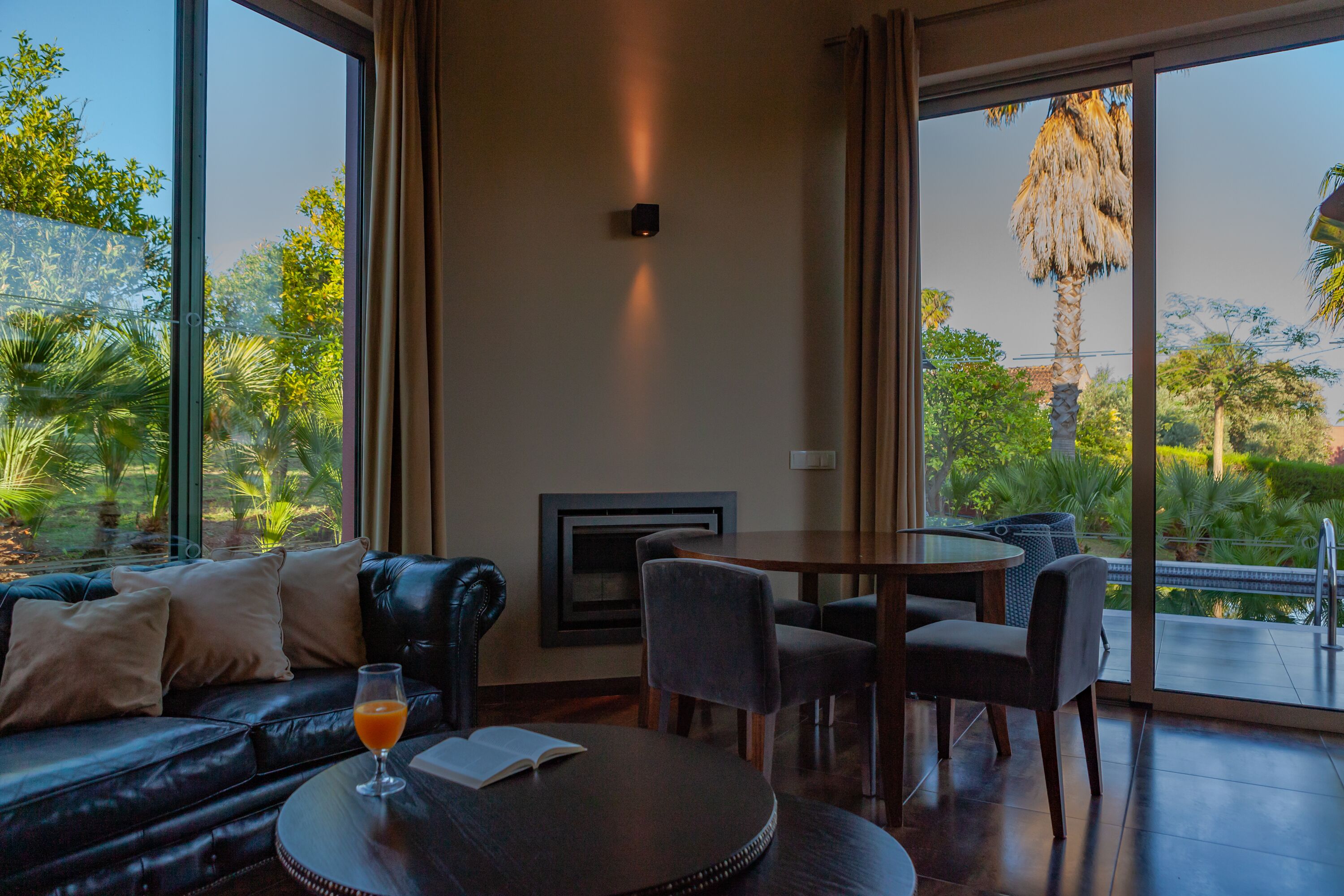 A modern living room with a leather sofa, a dining table with chairs, and a large window offering a view of greenery and a palm tree.