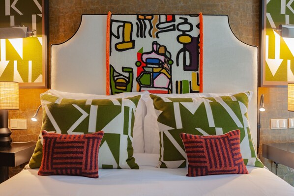 A bed with a geometric-patterned headboard and pillows, a bedside table with a lamp, and a wall with abstract art and light switches.
