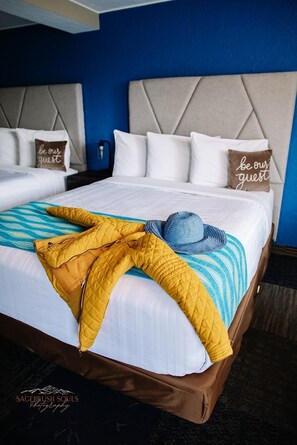 A hotel room with two beds, each with white bedding and a headboard. There are two pillows with the text "be our guest" on them. A yellow quilted blanket is draped over the bed. - American Classic Inn (Salida)