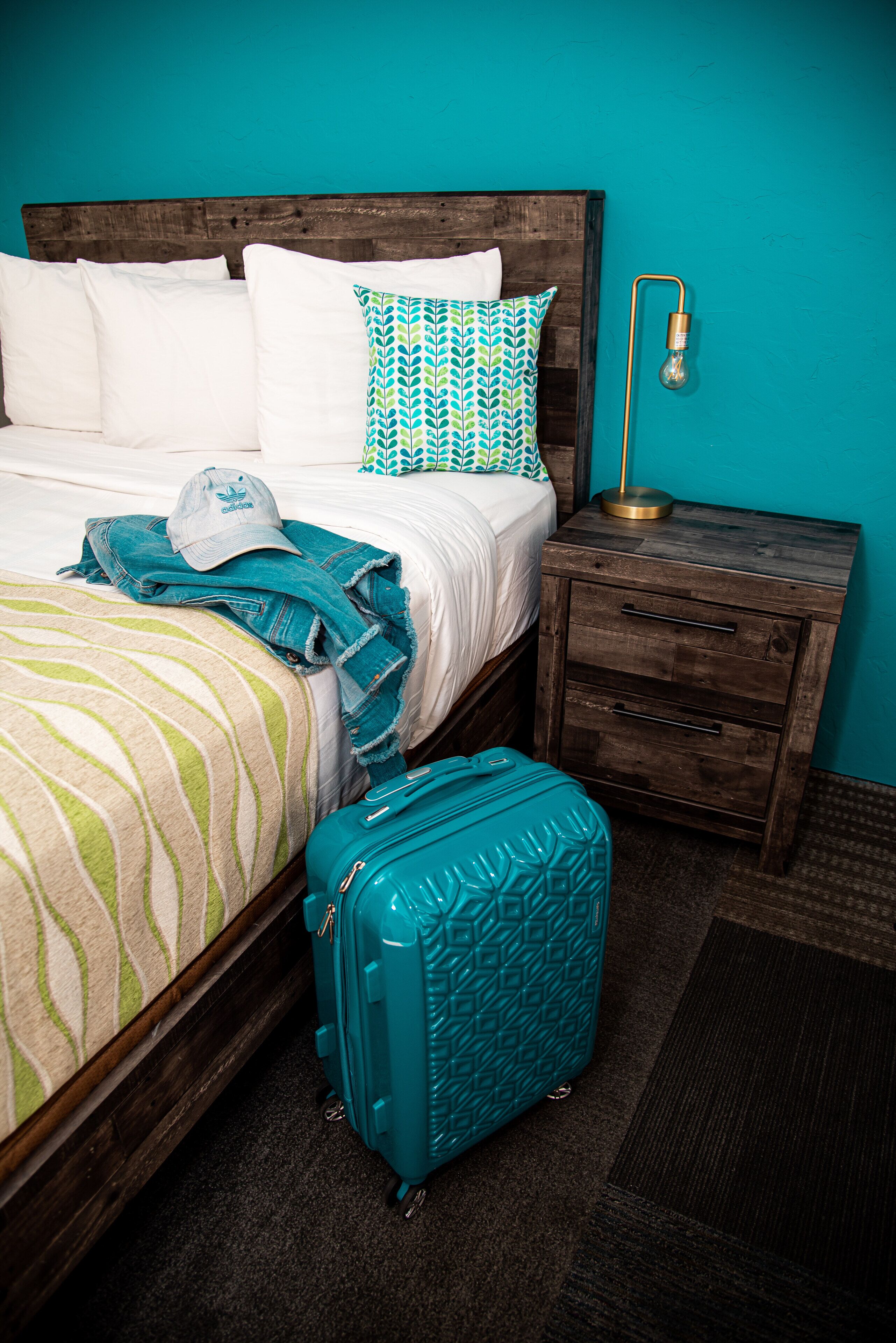 A bed with a striped blanket, a blue suitcase, and a wooden nightstand with a lamp.