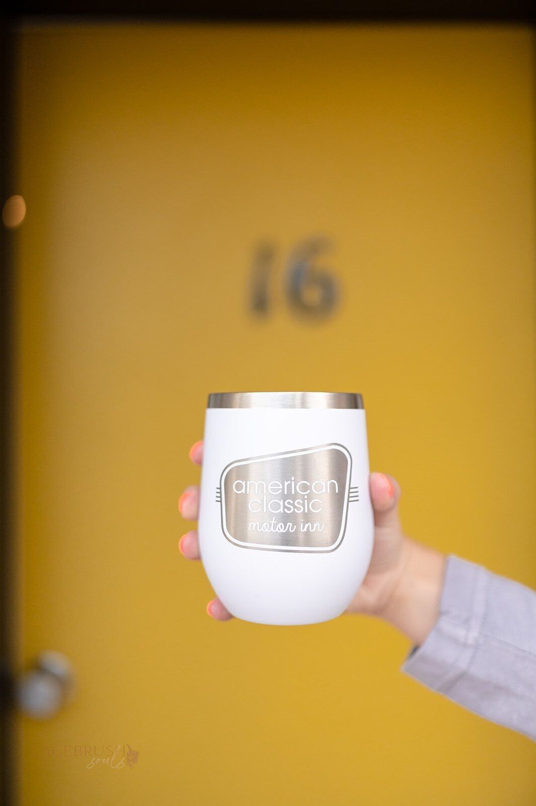 Una mano sostiene un vaso blanco con borde metálico y una etiqueta que dice 'American Classic Motor Inn'.