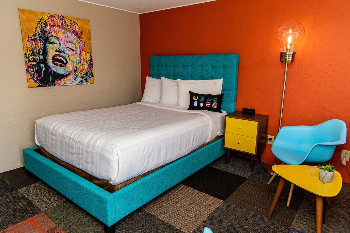 A bed with a teal headboard, white bedding, and a colorful painting above it. There's a yellow nightstand, a blue chair, and a floor lamp with a light bulb.