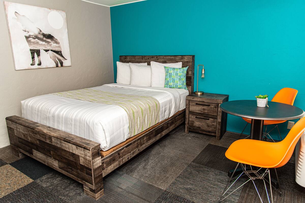 A bedroom with a wooden bed, a nightstand, a round table with an orange chair, and a teal wall.
