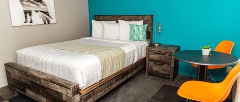 Un dormitorio con una cama de madera, una mesita de noche, una mesa redonda con una silla naranja y una pared verde azulado.