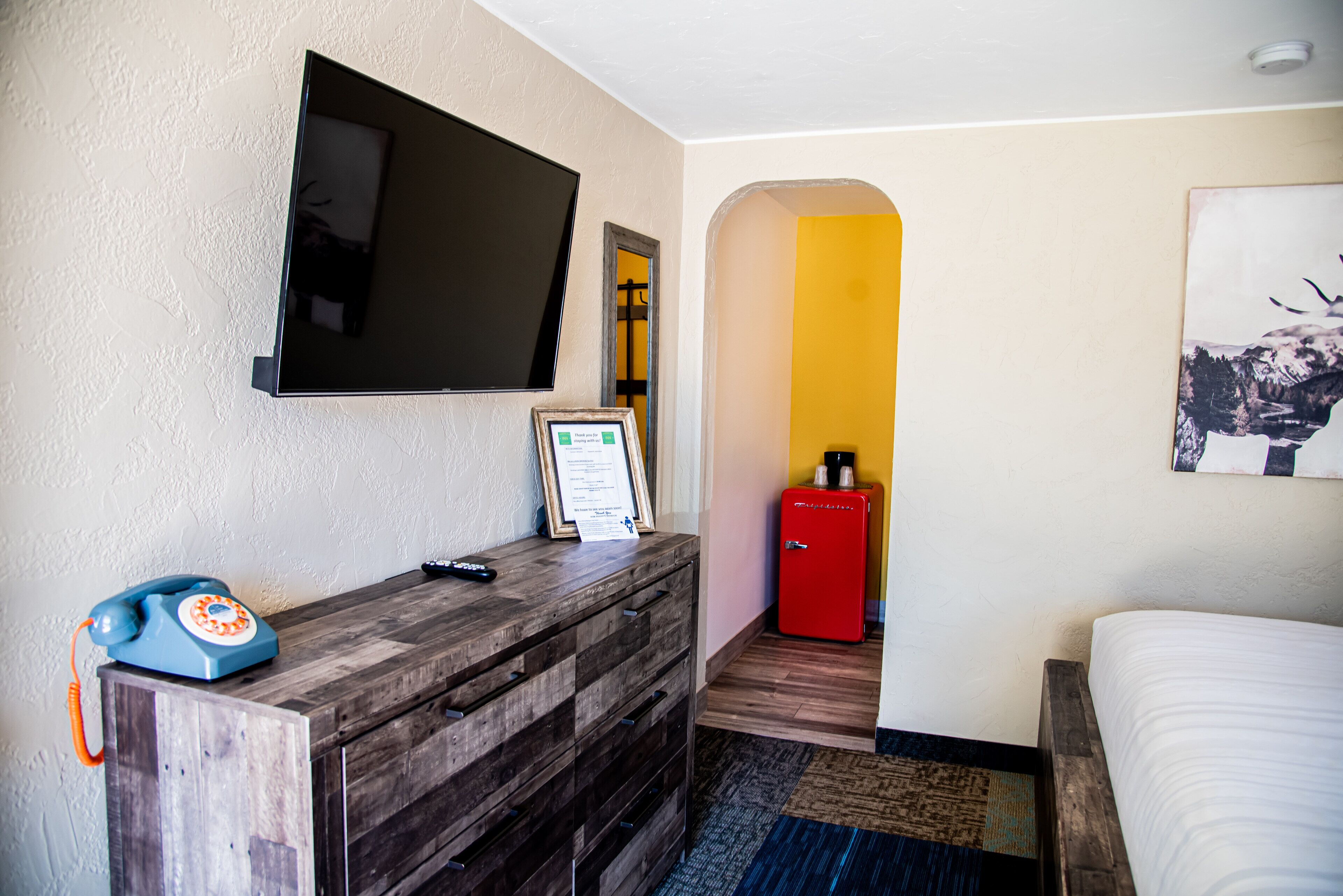 A hotel room with a wooden dresser, a television mounted on the wall, a red refrigerator, and a bed with white bedding.