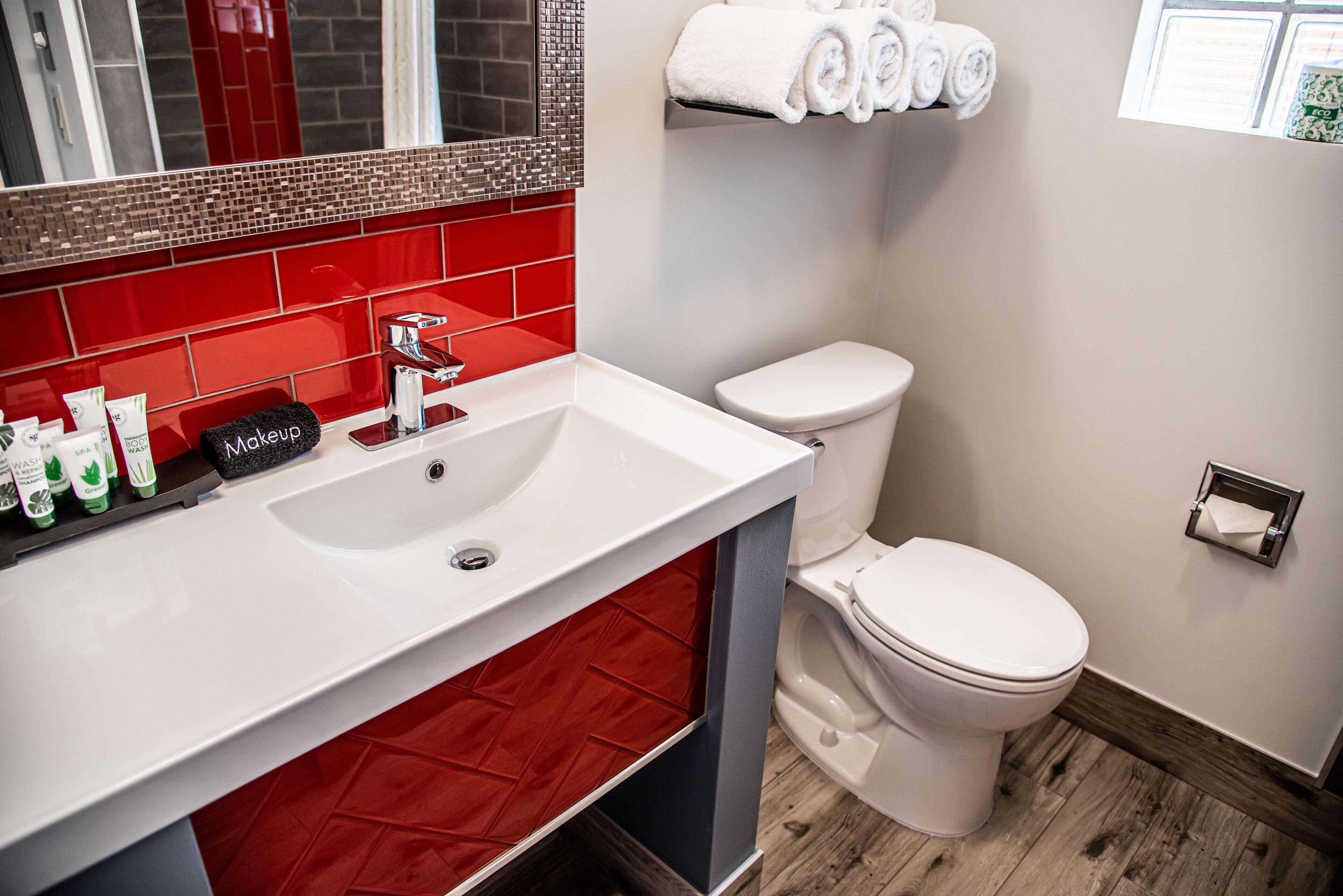 A bathroom with a white sink, red-tiled backsplash, a white toilet, and a towel rack with rolled towels.