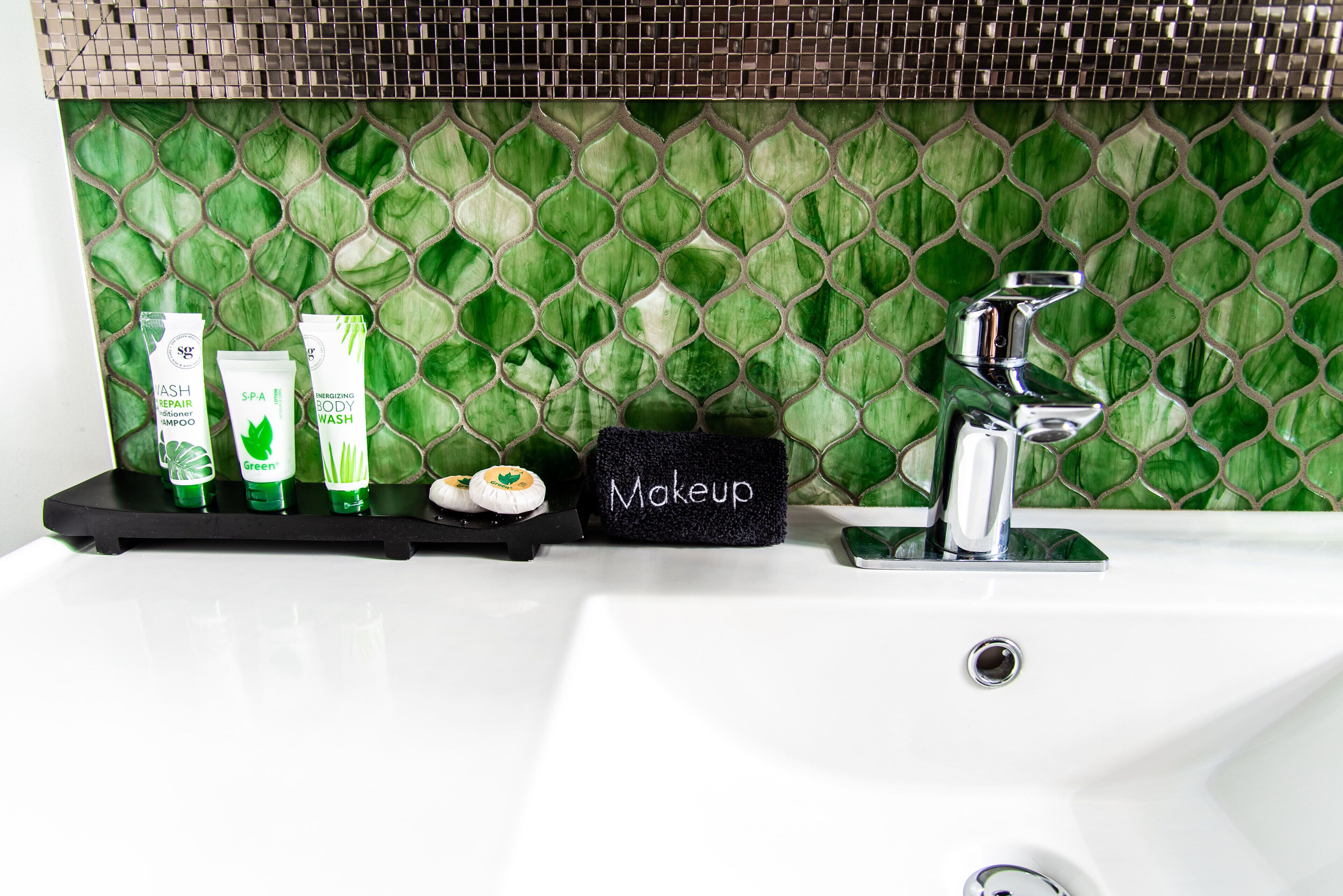 A bathroom vanity with a green mosaic backsplash, a black soap dish labeled "Makeup," and three green bottles of shampoo and conditioner.