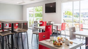 A modern dining area with tables and chairs, a flat-screen TV on the wall, and large windows. - B&B HOTEL Romilly-sur-Seine (Maizieres-la-Grande-Paroisse)