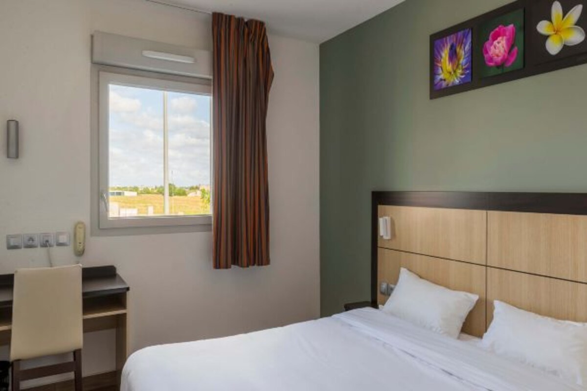 A hotel room with a bed, a desk, and a window with curtains. There are three paintings on the wall.