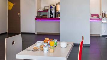 A breakfast table with various food items and drinks, a coffee machine, and a water dispenser in the background.