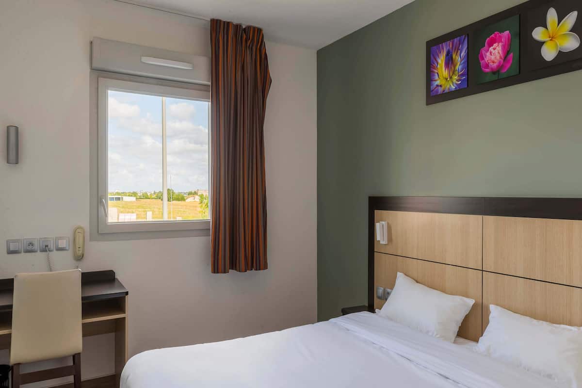 A hotel room with a bed, desk, chair, window, and artwork on the wall.