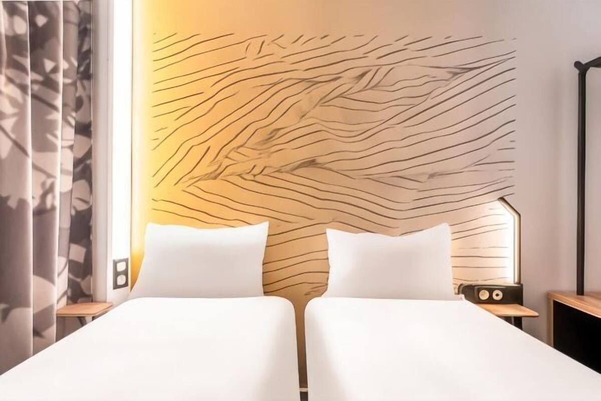 A hotel room with a modern design, featuring a large bed with white linens, a unique textured wall behind the bed, and a small bedside table with a radio.