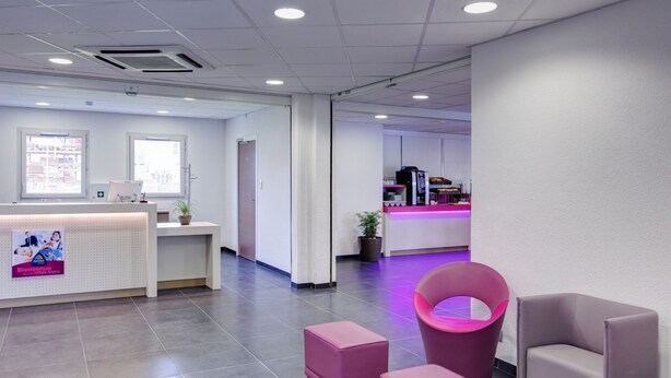 A modern reception area with a white reception desk, a pink chair, and a seating area with a pink ottoman. - B&B HOTEL Romilly-sur-Seine (Maizieres-la-Grande-Paroisse)