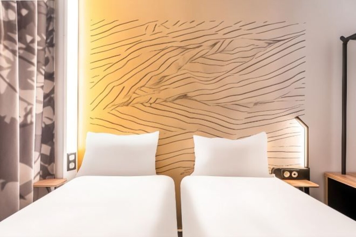 A hotel room with a modern design, featuring a large bed with white linens, a unique textured wall behind the bed, and a small bedside table with a radio.