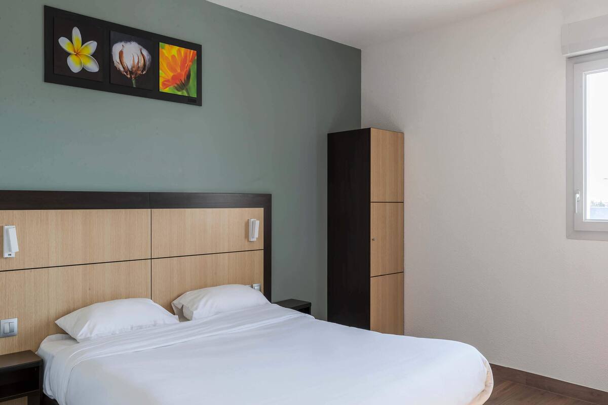 A neatly made bed in a hotel room with a wooden headboard, a small bedside table, and a wardrobe.