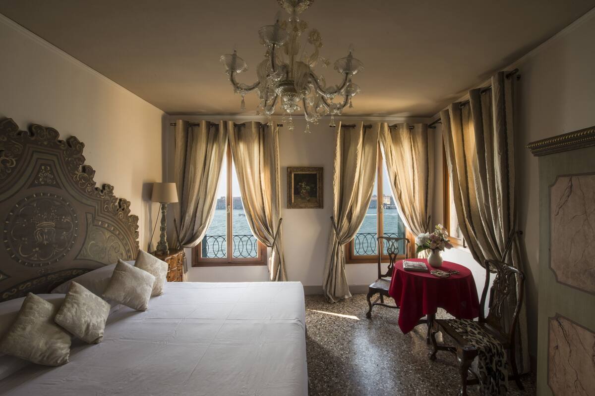 A hotel room with a large bed, a chandelier, a table with a red tablecloth, and a view of the sea through the windows.