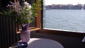 Ett fönster med utsikt över en kanal och byggnader, en vas med blommor och ett litet bord med en korg.