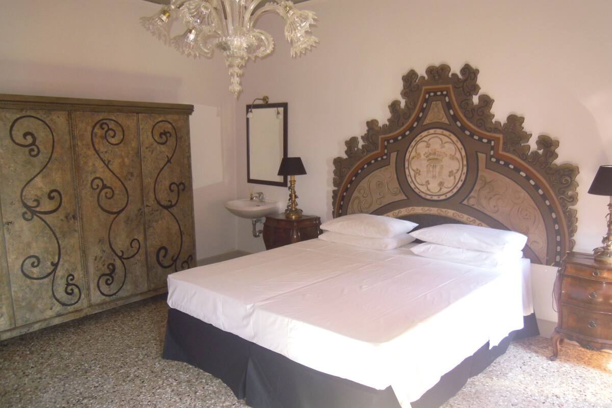A bedroom with a large bed, a wooden wardrobe, a nightstand with a lamp, and a decorative ceiling light.