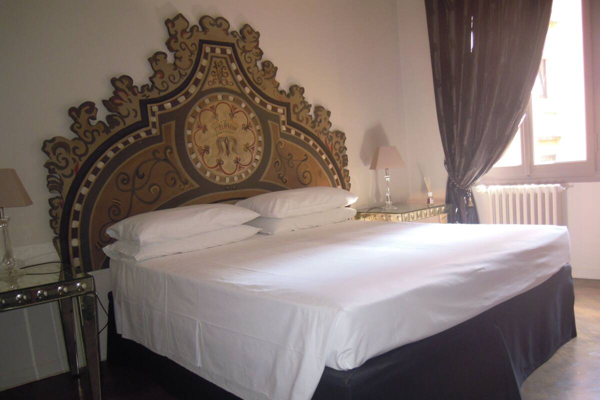A large bed with a decorative headboard, two bedside tables with lamps, and a window with curtains.