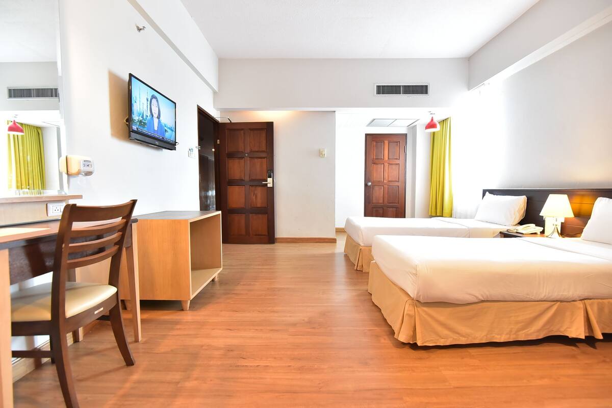 A hotel room with a wooden desk, two beds, a TV, and a door leading to another room.
